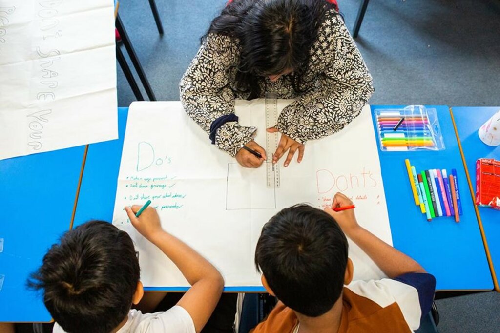 Children drawing the do's and don'ts of online safety with lots of colours on a large white paper on a sky blue table.